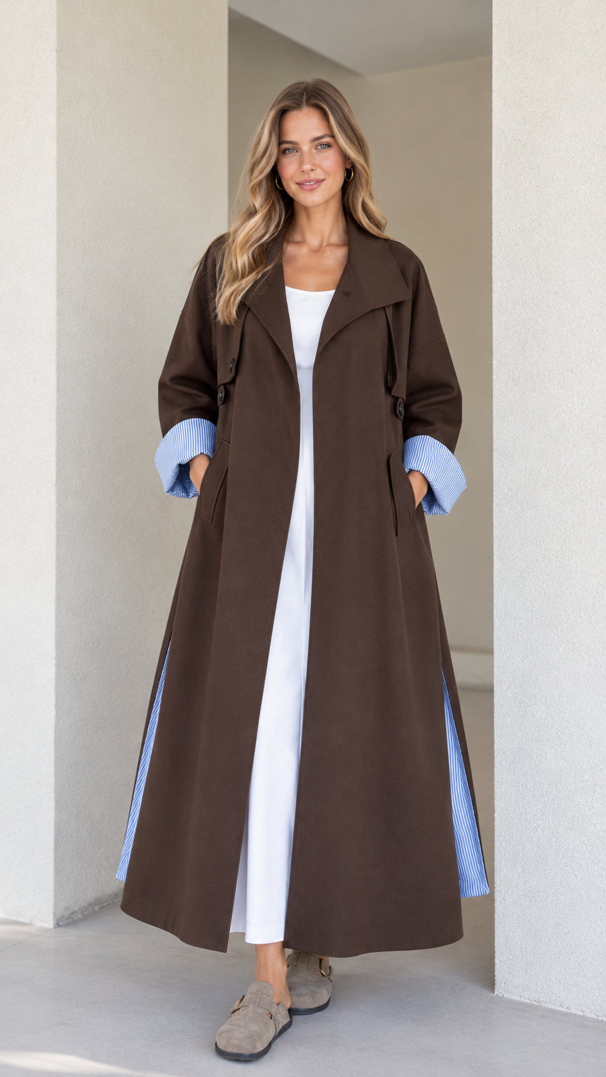 Front view of model wearing a brown maxi trench coat with striped turn-up cuffs over a white dress